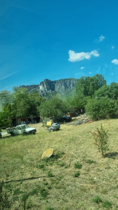 The image depicts a rural scene with a rugged, rocky mountain range in the background. In the foreground, there is a grassy field with several abandoned vehicles and structures. A light-colored pickup truck is parked on the left, with a smaller, grey vehicle, possibly a trailer or a small car, to its right. A yellow object, potentially a piece of machinery or equipment, is also visible in the field. Several trees and bushes are scattered throughout the landscape, and a rustic wooden shed or building is situated amidst the foliage. The sky is bright blue with a few scattered white clouds. The overall impression is one of a somewhat derelict, outdoor storage or workshop area set against a dramatic natural backdrop. This location is in Kalambaka, Greece, a town renowned for its proximity to the Meteora monasteries, which are built atop these distinctive rock formations.