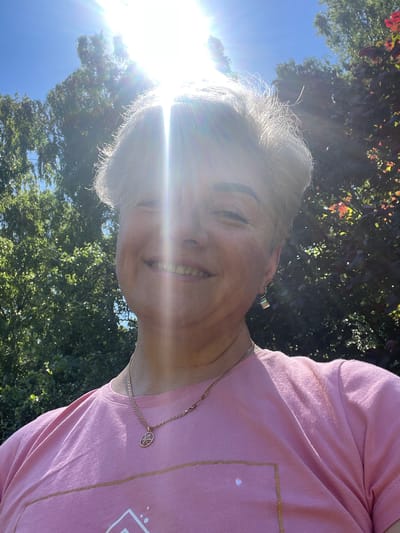 A woman with short, gray hair and a pink t-shirt smiles at the camera. She is wearing a necklace and earrings. The sun is shining brightly, creating lens flare. Trees and a blue sky are visible in the background. The location is identified as the city of Pervomaysky, Russia.