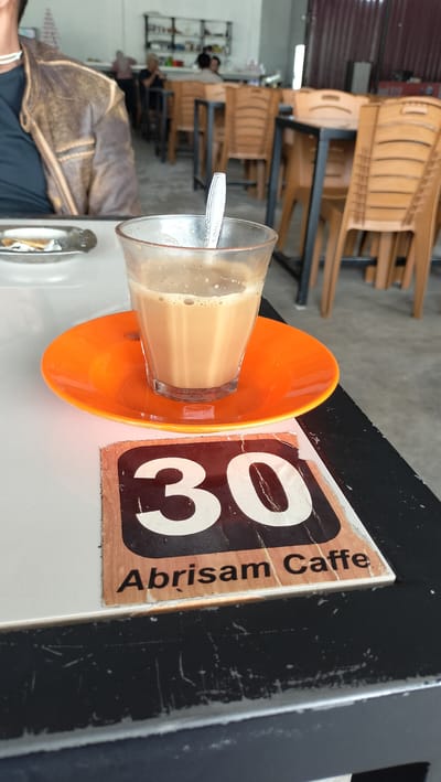 A table with a glass of coffee and a saucer is in the foreground of an Indonesian cafe. The table is black and white. A brown leather jacket is on the left side of the frame. In the background, there are several tables and chairs, and a shelf with various items. The overall setting is that of a casual dining establishment.