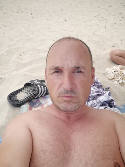 The image is a selfie taken on a sandy beach, likely in Osenovo, Bulgaria. A shirtless man with fair skin and a shaved head is in the foreground, facing the camera. He has short, graying hair on his head and stubble on his chin and upper lip. His skin appears slightly reddened on his neck and chest, suggesting sun exposure.

To his left, a black Adidas slide sandal lies on a towel. Behind him, a checkered shirt in purple and white is folded. To his right, a pile of small, white seashells is visible, and further back, the lower leg of another person can be seen. The background consists of undulating sand with small debris scattered across it. The lighting suggests it is daytime.