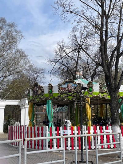The image depicts a colorful carousel in what is likely a park or public space in Urussu, Russia. The carousel is decorated with images of Disney characters. It is enclosed by a red and white striped fence. The scene is partially obscured by bare trees. The sky is overcast. Several individuals are standing near the carousel.