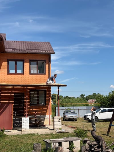 In Pavlovka, Russia, a man kneels on the edge of a partially constructed roof overhang of a two-story brick house. He is wearing a dark t-shirt, light-colored pants, and gloves. The house has a brown tiled roof and dark brown window frames. Below the overhang, there is a covered porch area with a red door and a window. In the yard, a young boy stands on a large piece of driftwood, looking up at the sky with his hand shielding his eyes. Two cars are parked in the grassy yard: a silver sedan and a white SUV. A metal fence runs along the background, behind which are trees and a partially visible rural structure. The sky is blue with scattered white clouds. The overall scene depicts outdoor construction and domestic life on a sunny day.