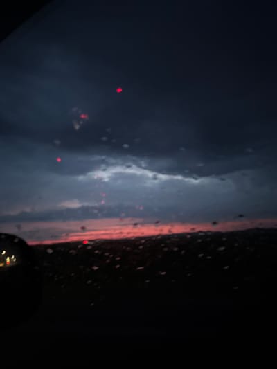 The image presents a forensic view from within a vehicle, looking outward through a rain-streaked window at twilight in Лабжанскі сельскі Савет, Belarus. The upper two-thirds of the frame are dominated by a heavily clouded, dark blue-grey sky. A distinct horizontal band of intense red and pink light stretches across the horizon, suggesting either sunset or sunrise. Numerous circular and elongated water droplets adhere to the glass surface, causing significant refraction and blurring of distant light sources. Scattered throughout the sky portion are multiple distinct, blurred, circular red light points, likely originating from distant artificial illumination refracted by the raindrops. The foreground in the lower-left quadrant shows a dark, curved object, possibly a vehicle's side mirror, partially obscuring the view, with a few indistinct, blurred yellow-white lights visible near its edge. The terrain beneath the horizon is predominantly dark and indistinct, appearing as a blurred, shadowy mass. No human figures or discernible structures are present in the distant landscape. The overall scene is characterized by low light, precipitation, and limited visibility.
