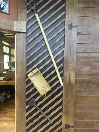 The image depicts a section of a rustic interior wall, likely indoors in Selyatino, Russia. The upper portion consists of vertically oriented wooden planks of varying widths, stained in a medium brown tone. Two black metal hinges are attached to the horizontal plank bordering the top of a panel below.
The panel below features a pattern of alternating diagonal wooden slats in natural and dark grey tones. Several tools are displayed on this panel. A long-handled pitchfork lies diagonally across the slats. To the right, a wooden tray with a small handle hangs by a string. Further to the right is a short-handled spade or shovel.
Below the panel is a wooden beam. The image provides a glimpse into the room below. This room appears to be a storage or utility area with a window and various objects.