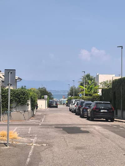 The image depicts a street in Sirmione, Italy, with a line of parked cars on the right side of the road. The street is paved with asphalt and marked with white lines. On the left, there is a building with a white fence and green hedges. In the background, a body of water, likely Lake Garda, is visible with distant hills and a clear blue sky. Several streetlights are positioned along the right side of the road. The scene appears to be a sunny day.