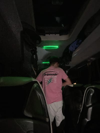 The image appears to be taken inside a vehicle, possibly a bus or large truck, at night. A person wearing a pink shirt and white pants is lying down, seemingly asleep. The vehicle's interior is dark, with some green lighting visible. There are seats and other vehicular components in the frame. The exact location in Lesichovo, Bulgaria cannot be determined from the image alone.