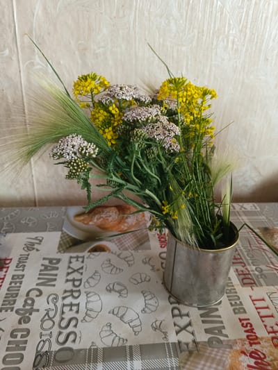 A still life composition features a bouquet of wildflowers and grasses in a metal vase, placed on a patterned tablecloth. The bouquet includes small yellow flowers, clusters of delicate pinkish-white flowers, and feathery green grass stalks. The vase is cylindrical with a brushed metal finish. The tablecloth displays text and illustrations of croissants, with a checkered pattern in sections. In the background, a textured wall with a faint pattern is visible. The scene is lit from the front, casting soft shadows. The overall impression is one of rustic simplicity. There is no indication of Kamenno-Angarsk, Russia, or any specific city or geographical location.