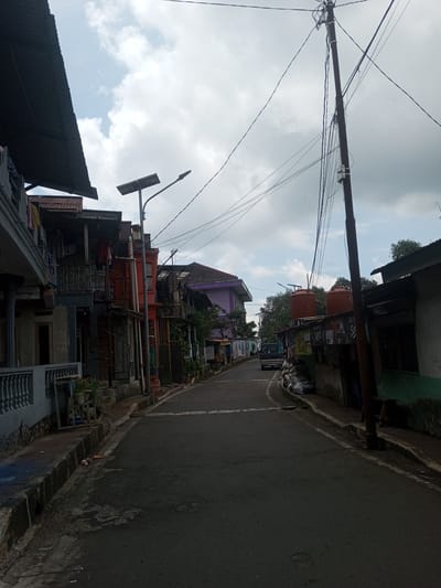 A narrow street in Ban Phouviengnoy, Laos, is depicted under a cloudy sky. The street stretches into the distance, with a dark-colored vehicle visible towards the end. Along the sides of the street, there are a variety of buildings, some with balconies and clothes hanging. One building is painted orange, and another is purple. There are utility poles with multiple wires running overhead. Several water tanks are visible atop one of the buildings. The pavement is asphalt, showing signs of wear.