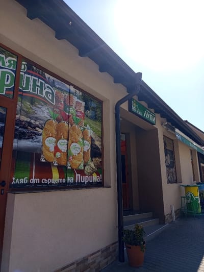 The scene depicts the sunlit exterior of a commercial building in Delchevo, Bulgaria. The building's facade is primarily beige stucco, with a dark brown gutter system and eaves. On the left, a large display window framed in dark brown features an advertisement for "Пирина" (Pirin) bread, showing several packaged loaves against a backdrop of a river and mountains. Text below the bread reads "Хляб от сърцето на Пирина!". The base of the building below this window is clad in stone-patterned tiles.
A dark brown drainpipe runs vertically down the facade. To its right, a recessed entrance area with light grey steps leads to a dark-framed doorway. A terracotta pot containing green foliage is positioned on the paved ground near the drainpipe. Above the entrance, a green sign with white text displays "ЛУПИ". Further to the right, a green and white striped awning extends over another section of the building, beneath which outdoor seating is present, including a yellow and green barrel-shaped table and at least two green stools. The sky above is clear and bright blue, showing significant lens flare from the sun. No people are visible in the scene.