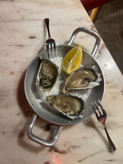 This image presents a close-up, overhead view of three raw oysters in their half-shells, served within a circular, two-handled aluminum pan. The oysters are embedded in a bed of coarse white salt that fills the pan. A single lemon wedge is positioned at the upper right within the pan. Two small, three-pronged metal oyster forks are visible: one is inserted prongs-down into the salt at the upper-center of the pan, and the other lies on the adjacent light-colored marble tabletop, extending from the bottom-right of the frame. The pan is centrally placed on the marble surface, which exhibits subtle light veining. Partial red elements, consistent with furniture or decor, are visible at the upper-right and lower-center edges of the image. This scene is observed within a commercial establishment in Caminha, Portugal.