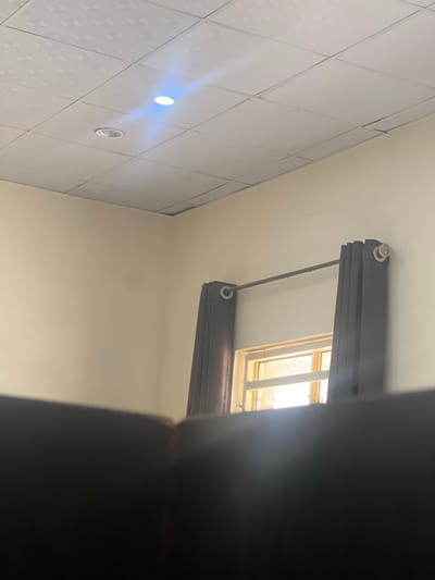 An indoor scene in Baganakwo, Nigeria, depicts a room with a white grid-patterned suspended ceiling. Two recessed circular light fixtures are present in the ceiling; one emits a bright blue-white light, while the other remains unlit. The walls are painted a light, uniform beige color. On the right side, a window is partially visible, equipped with dark brown or black grommet-top curtains hung on a silver-colored curtain rod. The curtains are drawn open to either side of the window. Bright natural light filters through the window, indicating daytime conditions. The lower foreground is occupied by a dark, out-of-focus object, likely a piece of furniture, obscuring the bottom portion of the wall and window.
