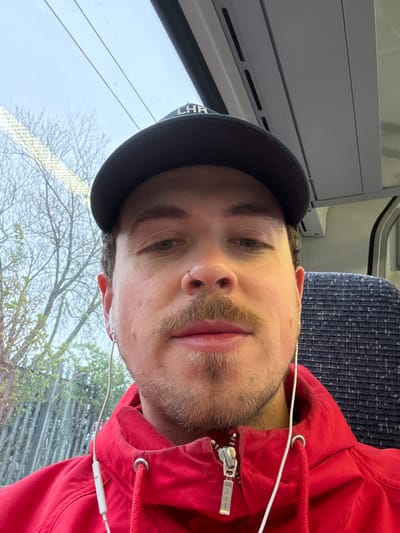 A male is seated on public transportation in Stevenage, UK, adjacent to a window. He is wearing a black baseball cap and a red hooded sweatshirt with a zipper pull reading "Aimé Leon Dore." White earbuds are visible. Outside the window, bare trees can be seen, suggesting a late autumn or early spring season. The seat upholstery is a patterned blue fabric.