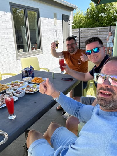 The image depicts a group of four men enjoying food and drinks outdoors. The location appears to be a patio or yard, likely in a residential area. Based on the surrounding architecture and landscaping, it's possible this scene takes place in Bredene, Belgium. Three of the men are visible from the waist up, while the fourth is seen from the knees up, with his face appearing close to the camera. The men are sitting around a dark-colored table laden with various snacks in white serving dishes. Some plates seem to contain meat dishes or appetizers, while one holds cheese-flavored snacks. Several cans of Red Bull energy drink and glass beverages in clear glasses filled with a red liquid, possibly juice or cocktails, are also present. One of the men is holding a bottle of Corona beer, and another one holds a glass with a reddish drink. All the men wear casual clothing. The man closest to the camera sports a light blue sweater and matching shorts, along with reflective sunglasses. One of the men poses with a thumbs-up gesture. A small child can be seen in the background. The weather appears to be sunny, casting distinct shadows. In the background, a white building with grey window frames is visible. The patio area features yellow chairs and a dark table surface, providing a stark contrast to the bright surroundings. A patch of green foliage adds a touch of natural color to the scene.