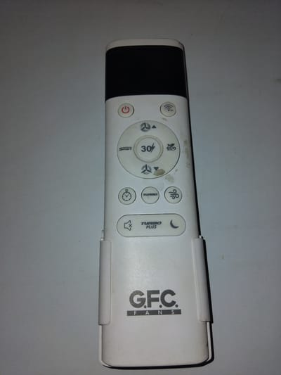 A white remote control with a black top is lying on a gray surface, possibly a table. The remote has various buttons with icons and text. At the top, there is a power button icon. Below that are buttons with icons representing a fan and "RF". Further down, there are buttons labeled "Smart", "30", and an "eco" icon. There are also buttons with fan speed indicators, a clock icon, a "TURBO" button, and a button with a wavy lines icon. Near the bottom, there is a "TURBO PLUS" button and a crescent moon icon. The bottom of the remote displays the logo "G.F.C. FANS". The remote appears to be slightly dusty. There is no indication of the location being in Chhina Uttar, Pakistan.