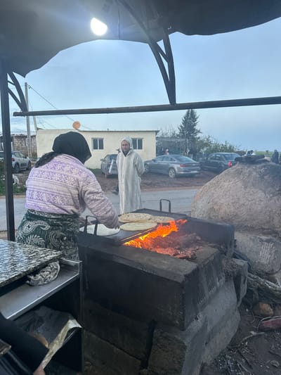 The image captures an outdoor scene featuring two individuals engaged in food preparation, likely street food, under an overcast sky. The overall setting suggests a rustic or informal environment, possibly a market or a roadside stall.

In the foreground, slightly to the left, a person is seen from behind, actively cooking flatbread on a large, flat metal griddle. This individual is wearing a light purple sweater with a white snowflake-like pattern, a dark head covering, and a patterned dark green skirt or long bottom. Their hands are visible, tending to the flatbread on the hot surface. There are several pieces of flatbread, resembling tortillas or similar unleavened bread, already on the griddle, with at least one being moved or placed.

The griddle is heated by a robust wood fire, with bright orange and yellow flames intensely visible beneath and around the cooking surface. The entire cooking station appears to be constructed from dark, rugged stone or concrete blocks, giving it a sturdy, traditional appearance. To the right of the cooking area, there's another section of the stall, possibly a counter or storage unit, with a marbled or speckled surface and some crumpled cloths or towels visible inside.

Standing opposite the cook, in the midground, is another person, facing towards the cooking activity. This individual is wearing a long, light-colored hooded robe or djellaba with a subtle pattern. Their gaze appears directed at the food being prepared. Their presence suggests they are either a customer, an assistant, or a companion.

Behind the two people, a paved or dirt road is visible, with several parked cars (sedans) lining it. Further in the background, there's a light-colored, two-story building with multiple windows, likely a residential or commercial structure. Beyond the building and cars, some sparse trees and distant landscape elements are visible. The sky is bright but overcast, suggesting either early morning, late afternoon, or a generally cloudy day, with a soft, diffused light.

To the far left of the frame, part of a dark, tent-like canopy structure is visible, under which the cooking is taking place. A bright, bare light bulb or lantern is hanging from this structure, providing additional illumination, hinting that it might be dusk or dawn, or simply a consistently lit stall. Large, natural-looking boulders or rocks are present in the far right background, blending into the rustic aesthetic of the scene.

The image depicts a vibrant, informal cooking operation, centered around the preparation of fresh flatbread over an open fire, a common sight in many cultures. No discernible text is visible in the image.