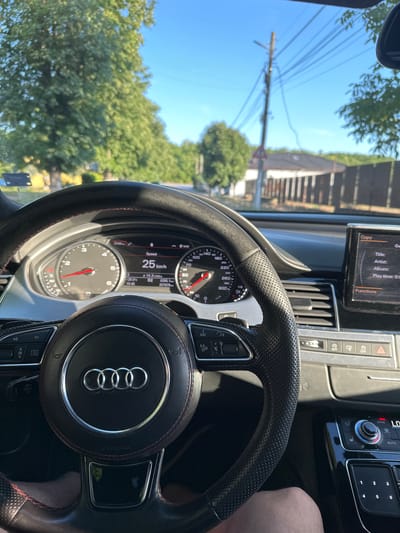 This is a close-up, interior shot of the driver's perspective from inside an Audi car. The car is moving at 25 km/h, indicated on the dashboard. The driver's legs are visible at the bottom of the frame. Outside the car, a residential area with houses and trees is visible under a clear blue sky. The scene appears to be in Marten, Bulgaria, given the context. The car's infotainment system displays music information and other vehicle functions.