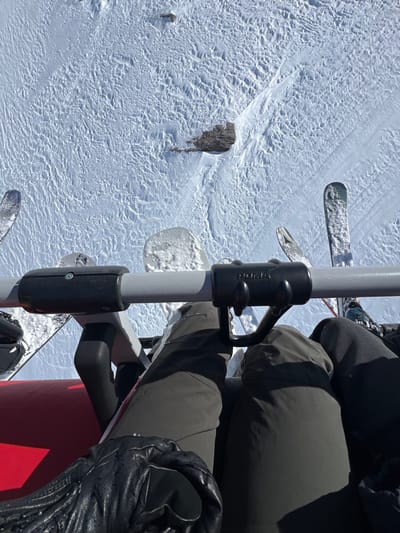 The image captures a close-up view from within a ski lift cabin, looking down towards the snowy slope below. The cabin is occupied by a single individual, whose lower legs and feet are visible. Their right leg is extended, clad in dark grey ski pants, and their left leg is bent at the knee, with their foot resting on the lift's metal bar. They are wearing a pair of black ski gloves, one of which is visible on their left hand. The scene is set against a backdrop of a snow-covered slope, with the texture of the freshly fallen snow clearly visible. The coordinates provided (latitude: 45.421, longitude: 7.013) pinpoint the location as Val-d'Isère, France. There are two pairs of skis secured to the lift's frame, indicating the individual is a skier. The image was likely captured during a sunny winter day as the bright white snow reflects the light. The overall atmosphere is one of tranquility and quietude, typical of a winter ski resort.