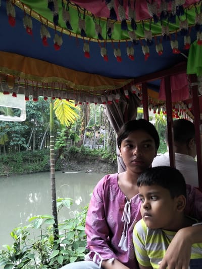 The image shows two children sitting inside a small boat or carriage, likely a type of traditional water transport, in what appears to be Southkhali, Bangladesh, given the environment.

The older child, a teenage girl, is wearing a purple shirt with white embroidery and white decorative tassels at the neckline. The younger child, a boy, is wearing a striped green and yellow shirt. He is seated close to the girl, and her arm appears to be around him. They both look off to the side, possibly at the passing scenery. Another person, partially visible, is behind them, wearing a white shirt.

The boat's interior is decorated with colorful fabrics in shades of blue, green, pink, and yellow. Hanging from the fabric are decorative tassels, which appear to be a mix of white and red.

Outside the boat, there's a canal or river with greenery along the banks. Trees and foliage are visible in the background, typical of a tropical or subtropical environment. A palm tree is directly in view. The water is somewhat murky in color. Plants are visible in the foreground at the bottom of the image.