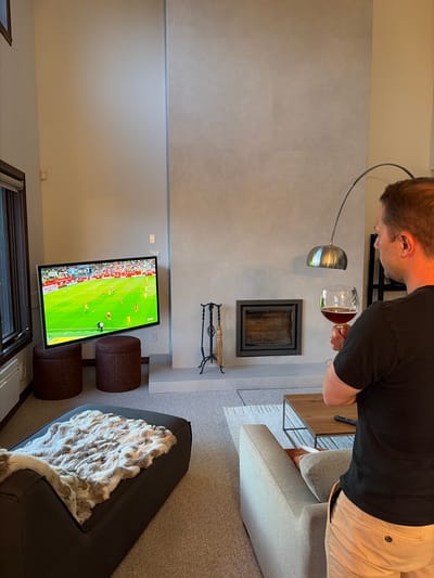 In a contemporary living room, a man in a black t-shirt and khaki shorts stands facing away from the viewer, holding a wine glass filled with red liquid. He is positioned to the right of a large, modern fireplace with a metallic insert. A sleek, arc-shaped floor lamp with a silver shade extends over his head. To the left of the fireplace, a large television displays a soccer match. Two dark brown, cylindrical ottomans are placed in front of the television. A dark grey chaise lounge with a furry throw is visible in the foreground, on the left side of the image. A light-colored sofa and a wooden coffee table with a remote control are in the mid-ground, to the right. The room has light-colored walls and carpeted floors. The overall ambiance suggests a relaxed evening at home, likely in a ski resort town like Mont-Tremblant, Canada, known for its modern architecture and recreational lifestyle.