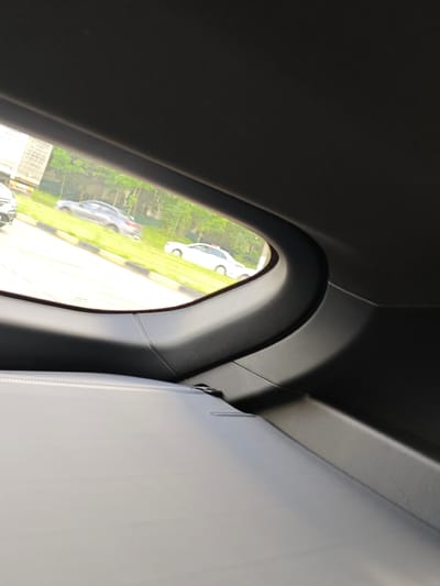 The image captures a view from inside a vehicle, looking out of the rear window. The window frame, made of dark grey plastic, curves upwards and to the right. Below the window, a light grey, textured surface, likely part of the car's interior, takes up the lower portion of the frame.
Through the window, a street scene is visible. On the left side of the road, a grey sedan is parked, and beyond it, a large building with visible windows and possibly balconies is partially obscured by trees. The road itself is paved, and a grassy verge runs alongside it. Further down the road, a white sedan is in motion. Lush green trees line the background, suggesting a setting with ample vegetation. The lighting indicates it is daytime, with bright sunlight casting shadows.
The context provided places the location in Krasnenskoye village, Belarus. No people are directly visible in the image. The scene depicts a typical roadside view from a car, suggesting a moment of travel or waiting. There is no visible text in the image.