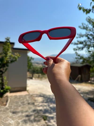 A hand, holding a pair of red-framed sunglasses up to the sky, is the main subject of this outdoor photograph. The sunglasses, with dark lenses, are positioned in the foreground, obscuring the upper portion of the hand and arm. The background features a clear blue sky, with a few blurred green tree branches visible. Further back, a light-colored building with a dark roof is partially visible on the left, and a small wooden structure sits in the middle ground on the right. The terrain appears to be an outdoor area with stone paving in the foreground and a sandy or gravel path beyond. The scene likely takes place in Cannara, Italy, given the rural landscape suggested by the vegetation and the presence of structures. The lighting indicates it is daytime, likely sunny, given the shadows and the bright sky.