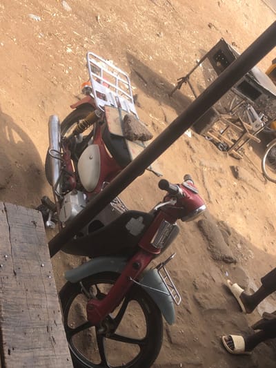 The image displays an outdoor scene in Unguwar Fatika, Nigeria, dominated by dry, uneven brown earth under direct sunlight. A red and light blue/grey Honda Cub-style motorcycle is stationary, angled from the lower left to the upper right. The motorcycle features a silver exhaust pipe on its left side, a silver rear luggage rack to which a cardboard sheet and a dark, textured bundle are secured, and a worn black seat. A dark, diagonal metal bar extends across the mid-ground. To the right of the bar, a rudimentary metal cart or stand and a partial bicycle wheel are visible. In the lower right foreground, two dark-skinned feet wearing light-colored open-toed sandals are present, indicating an individual standing nearby. Rough wooden planks are visible in the lower left foreground, suggesting an elevated viewpoint.