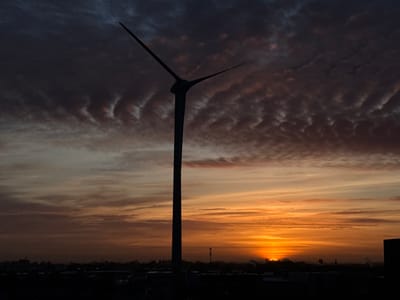 A prominent wind turbine stands in stark silhouette against a dramatic sunrise or sunset sky over Maasdijk, Netherlands. The central turbine, with its three distinct blades, dominates the frame, appearing static. The sky presents a multi-layered spectacle: the upper left portion features dark, textured clouds with visible undulations, possibly mammatus formations, in shades of dark blue and grey. Below these, the sky transitions into warm hues of orange, yellow, and soft pink as it approaches the horizon.
The sun is partially visible as a bright, glowing orb, peeking just above the distant, silhouetted landscape. This distant horizon reveals a low-lying urban or industrial area, with faint outlines of buildings, trees, and what appears to be a communications tower. The foreground, like the turbine and distant land, is also rendered in deep shadow, emphasizing the light of the sky. The overall impression is one of serene power and natural beauty, captured during the golden hour. No visible text is present in the image.