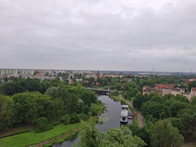 This is an aerial, eye-level, medium-shot photograph of the city of Pila, Poland, on an overcast day. The scene depicts a river flowing through a verdant urban landscape. Lush green trees dominate the foreground and midground, lining the riverbanks and interspersed with residential buildings. A concrete structure, possibly a house or dock, is situated on the river. A bridge spans the river in the midground, with buildings and more trees visible on either side. The background features a dense collection of urban buildings, including apartment blocks and older structures, extending to the horizon. Industrial elements, such as smokestacks and a crane, are visible in the far distance. The sky is uniformly gray and cloudy, suggesting a cloudy or possibly rainy day. There is no visible text in the image. The overall impression is of a tranquil, somewhat subdued urban environment integrated with nature.