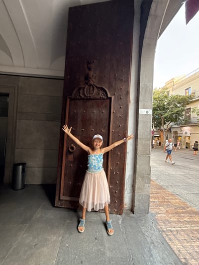 A young girl wearing a blue and white floral top, a light pink skirt, a white cap, and blue sandals stands with her arms outstretched in front of a large, dark wooden door. The girl is positioned slightly to the left of the door, which appears to be part of an old building. The door is ornate, with decorative carvings at the top and numerous dark studs. To the left of the door, a dark grey trash can is visible. The ground is paved with dark grey stone. To the right of the girl and the door, a concrete archway reveals a street scene. A man in a grey t-shirt and blue shorts walks away from the camera on the right side of the street, and other people are visible further down the street in front of shops. The street is paved with a mix of grey and reddish-brown wooden planks. A sign with the number "112" is mounted on the concrete pillar of the archway. The architecture suggests a historical urban setting in Mataró, Spain. The lighting indicates it is daytime, possibly late afternoon.