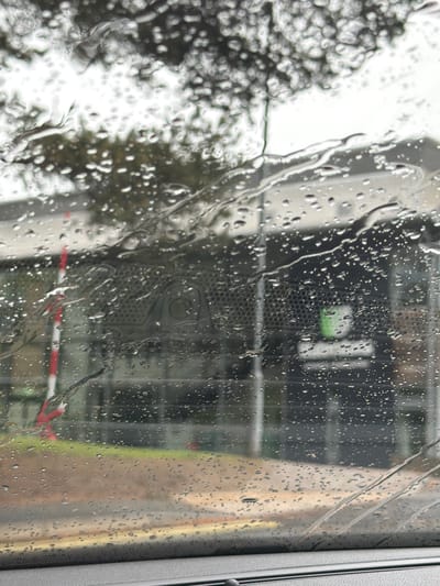 The image shows a view through a rain-streaked window. Outside, indistinct buildings and trees can be seen. There appears to be a red and white striped traffic barrier in the background. A dark object with a perforated surface is partially visible through the raindrops. Based on the composition and context, it's impossible to confirm if this location is in Durham, United Kingdom, without additional information.