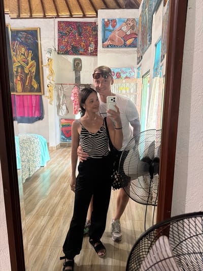 A full-body mirror selfie shows a Caucasian man and an Asian woman standing close together in a room with numerous paintings on the walls. The man, on the right, wears a white t-shirt and sunglasses, holding his phone to take the photo. The woman, on the left, is in a black and white striped camisole and black pants, with his arm around her waist. The room appears to be an art gallery or shop, with paintings depicting various scenes, including abstract figures and a nude woman. There is a bed with a blue and white patterned cover on the left side of the image, and a black standing fan is visible on the right. The floor is tiled with a light wood-grain pattern. The context suggests the location is Tejakula, Indonesia.