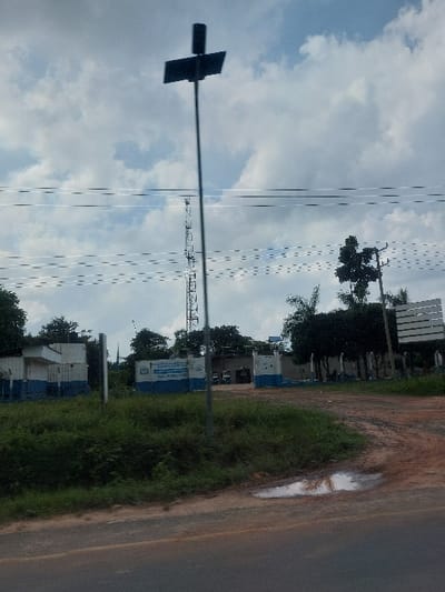 The image displays a roadside scene in Kisanga, Tanzania, under a cloudy sky. In the immediate foreground, a dark grey asphalt road surface is visible. To the right, a reddish-brown dirt path curves into a compound, containing a small, irregularly shaped puddle reflecting the sky. Green, unkempt grass and low-lying vegetation line the road and the edge of the dirt path.
A tall, slender metallic pole stands prominently in the midground, featuring a dark, rectangular solar panel oriented towards the sky, and a cylindrical object mounted at its apex. Behind this pole, multiple thin, white overhead power lines extend horizontally across the frame. Further to the right, a dark wooden utility pole with crossarms supports additional electrical lines.
The background reveals a walled compound with buildings painted white and blue. A prominent sign on the central wall of a building is partially legible, displaying "KITUO CHA POLISI" (Police Station). Several vehicles, including what appear to be cars or SUVs, are parked within the compound, visible through an open entrance or gate area. Dense green trees and foliage are scattered within and around the compound. A tall, metallic lattice communication tower is visible to the left, standing behind the compound structures. The sky above is largely overcast with white and grey clouds. No individuals or specific actions are discernible.