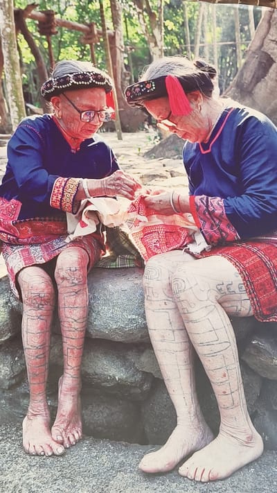 The image depicts two elderly women performing needlework outdoors in Jiyang District, China. Both individuals are seated on large, irregular grey rocks, facing slightly towards each other and intently focused on fabric held between them.
The woman on the left wears a dark blue long-sleeved shirt with embroidered red, orange, and gold cuffs, paired with a red patterned knee-length skirt. She has a dark, beaded headpiece, red-framed eyeglasses, and visible tattoos or markings on her face, particularly around her mouth and cheeks. Her legs, from above the knees down to her bare feet, are extensively covered in black linear and geometric tattoos, with some areas of skin appearing reddish. Her feet are aged and bare.
The woman on the right wears a similar dark blue long-sleeved shirt with patterned red and black cuffs, and a red patterned knee-length skirt. She sports a dark, embellished headpiece featuring a prominent red tassel on the side, and orange-framed eyeglasses. Her face also exhibits visible tattoos or markings. Her legs, like those of her companion, are extensively tattooed with black linear and geometric designs extending to her bare, aged feet.
Both women's hands are engaged with pieces of fabric; one piece is light-colored with red patterns, and another is primarily red, suggesting a shared activity of crafting or mending.
The immediate environment consists of the large grey rocks serving as seating. The background shows a blurred outdoor setting with green foliage and trees, indicative of a natural or wooded area. Vague light-colored wooden structures are discernible in the distant background among the trees. The scene is illuminated by natural daylight.