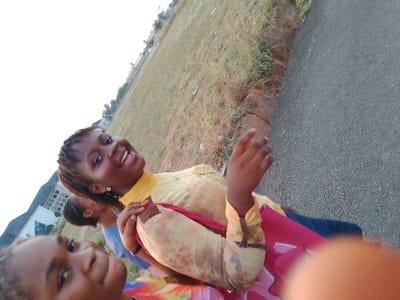 Three young women are standing on a gray asphalt road in Umueshi, Nigeria. The women are smiling and appear to be taking a selfie. Behind them is a grassy field. In the distance, there are buildings and a clear sky.