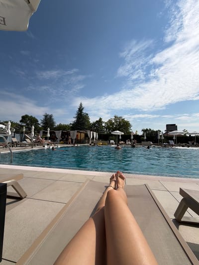 The image depicts a sun-drenched scene at a swimming pool, likely located in Hisarya, Bulgaria, given the context provided. In the foreground, a person's legs are stretched out on a reclined lounge chair, their feet visible with toes pointed upwards. The person is wearing a delicate ankle bracelet on their left ankle. Beyond the lounge chair, a large, clear blue swimming pool dominates the mid-ground. Several people are visible in the pool, some swimming and others standing. The poolside area is paved with light-colored tiles and lined with lounge chairs and umbrellas, suggesting a resort or hotel setting. In the background, lush green trees form a backdrop, and a covered structure with white curtains and yellow seating is visible near the pool's edge, possibly a bar or dining area. The sky above is a brilliant blue, dotted with scattered white clouds, indicating a clear and pleasant day. The overall atmosphere is one of relaxation and leisure.