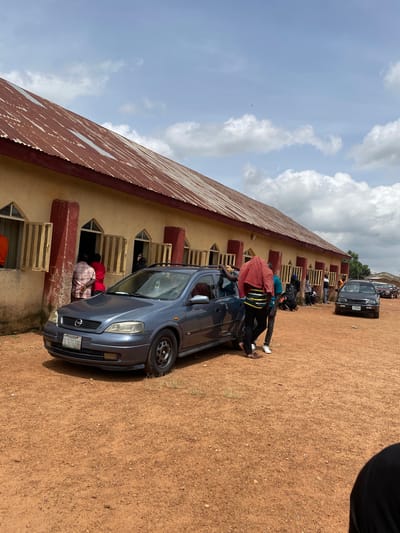 A long, light-yellow building with a rusty, corrugated iron roof stands under a cloudy sky. The building has several open windows with wooden bars. In front of the building, parked on a dirt ground, are several cars. A dark blue hatchback is in the foreground, facing right. Several people are standing near the cars and the building, some appearing to be talking. A black Volkswagen Golf is parked further down the dirt lot. The scene appears to be an outdoor gathering in Ban, Nigeria.