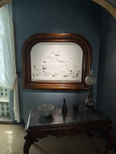 A dimly lit interior space presents a tableau of art and decorative objects. A table, its top marbled in grey and white, stands in the foreground. Atop it, from left to right, sit a shallow porcelain bowl, a small dark bottle, a miniature dark metal incense burner, and a lamp with a silvered base and a spherical white glass shade. Above the table hangs a framed bas-relief sculpture. The frame is of dark wood, arched at the top, and encloses a white-toned scene of numerous figures, including one prominent figure in the sky. To the left, a window is draped with a sheer white lace curtain, allowing a glimpse of a staircase outside. The wall is painted in a muted blue-grey. The scene appears to be within Quinta da Regaleira, a UNESCO World Heritage site in Sintra, Portugal, known for its opulent architecture and gardens. The bas-relief might represent a historical or mythological scene, characteristic of the estate's elaborate artistic program.