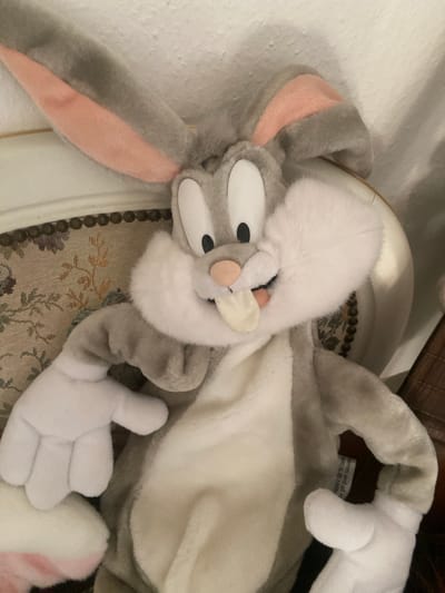 A plush Bugs Bunny toy is positioned on an upholstered surface, possibly a chair or sofa, with a detailed floral pattern visible beneath it. The toy, predominantly gray and white, features large white eyes with black pupils, pink inner ears, and an open mouth with a pink nose and a white tongue sticking out. Its hands are white and cartoonishly shaped. The background is mostly white, with a hint of textured wall and a wooden surface to the right. A tag is partially visible on the toy's body, showing the text "BROS. © 1999". The scene appears to be indoors. The specific location in Kassel, Germany, cannot be determined from the provided image.