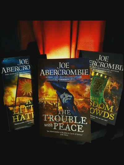 The image displays three Joe Abercrombie books arranged on a dark surface, possibly a table, with a red, illuminated backdrop. From left to right, the books are "A Little Hatred," "The Trouble With Peace," and "The Wisdom of Crowds." All books feature prominent author and title text. The lighting casts shadows and highlights, creating a dramatic effect. There are no people or indications of a specific city like Norcia, Italy, in the image.