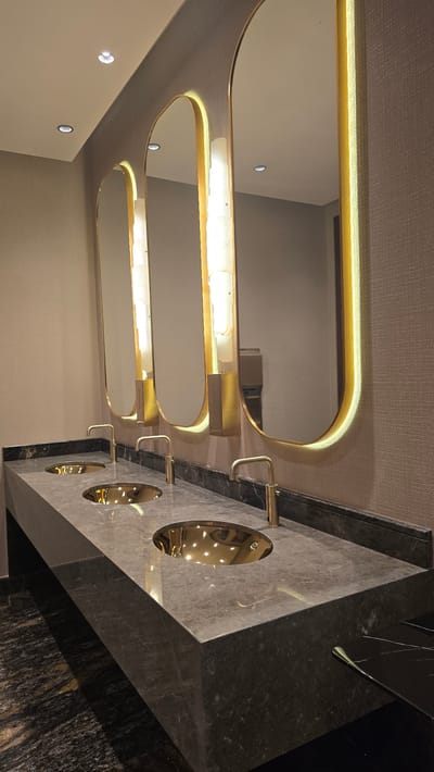 The image shows a row of three modern, oval-shaped sinks with gold faucets, set into a gray marble countertop. Each sink has a reflective gold basin. Above the sinks are three large, arched mirrors framed in gold, each with vertical, illuminated panels to the sides. The walls are covered in a textured, neutral-toned wallpaper. The lighting is warm and highlights the gold accents. The scene appears to be a well-maintained public restroom or vanity area. The location is identified as Agios Epiktitos, Northern Cyprus / Cyprus.