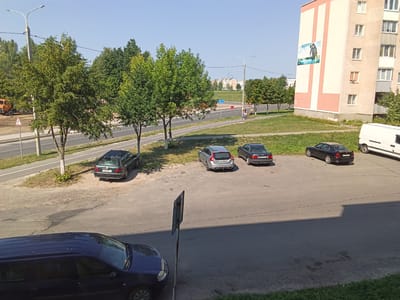 The image depicts a residential area in Drahichyn, Belarus, on a clear, sunny day. In the foreground, a dark blue station wagon is parked on the asphalt. Behind it, a grey station wagon and two dark sedans are parked in a lot. Further in the background, a white van is parked. A paved road runs along the side of the parking area, with a bicycle lane visible. Trees line the road, and a multi-story apartment building with a light pink and white facade stands to the right of the frame. A large mural is displayed on the side of the building. A street lamp is visible on the left side of the image. Two individuals, a man and a woman, are walking on the sidewalk beside the road. An orange construction vehicle is visible on the far left of the image.