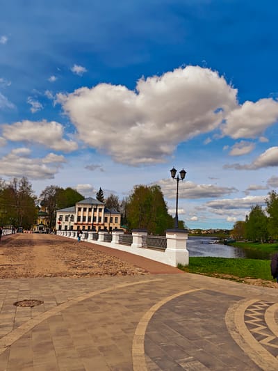 The image shows a view from a cobbled bridge in Uglich, Russia, under a partly cloudy sky. A prominent white cloud in the sky resembles a heart shape. A two-story historical building with a colonnaded facade stands to the left of the bridge. A black lamppost is located on the bridge near the river. The bridge has a white railing. There are people walking on the bridge and along the riverbank. The river flows to the right, with greenery along its banks. The foreground shows patterned paving stones.