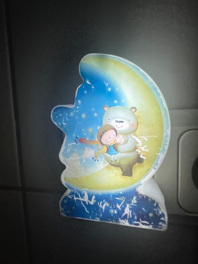 A close-up, side-view photograph shows a night light with a crescent moon and a bear hugging a child. The night light is shaped like a silhouette, with the moon and characters set against a starry sky. The moon is yellow and blue, and the sky is dark blue with white stars and streaks of white that could be clouds or abstract designs. The bear is light brown and hugs a child who is wearing a blue shirt. The child has brown hair and is smiling. The night light is placed against a dark background that appears to be a wall or cabinet. There is a faint, blurry grey object to the right of the night light. The image is grainy and the lighting is dim. The location is not specified in the image or provided in the prompt.