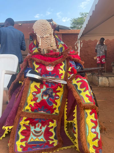 In Porto-Novo, Benin, a traditional Egungun masquerade takes place, featuring a costumed individual adorned in a vibrant, heavily embellished outfit. The costume is a profusion of colorful fabrics, primarily red and yellow, with intricate appliqués and fringed borders in shades of brown and gold. A striking headdress, constructed from numerous small white shells, crowns the figure, adding to its imposing presence. The masquerader holds a metallic object that appears to be a sword or blade, and a wooden staff. Standing behind the masquerader, a man in a denim shirt is visible. Further in the background, another young man wearing a patterned t-shirt and red shorts observes the scene, standing near a white tent structure. The ground is composed of reddish-brown earth, typical of the region. The backdrop features a building with an orange-brown textured wall. The sky above is clear and blue.