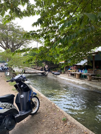 A vertical, medium shot captures a scene near a canal on a sunny day in Kresek, Indonesia. In the foreground, a black and blue scooter is parked on a dirt and gravel path. The scooter is angled to the left, with its front wheel and handlebars visible. The path leads to a concrete-lined canal filled with murky green water, reflecting the dappled sunlight filtering through overhead trees.

Beyond the canal, a small, open-air market or food stall is set up under a canopy of leaves. Several wooden tables and benches are arranged for seating. A few people, including at least one man and two women, are gathered around the tables, some appearing to be customers and others vendors. Coconuts are piled on one of the tables, suggesting fresh produce is being sold. In the background, more trees and foliage are visible, with a glimpse of buildings and a street with a few other motorcycles parked. The overall impression is a relaxed, local gathering spot by a waterway.