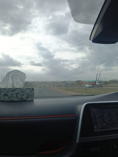The image shows a view from inside a car on a road in Athi River ward, Kenya. The dashboard is visible, with a tissue box on the left and a car display screen on the right. The road stretches ahead, with several vehicles including a large truck, a white SUV, and some smaller vehicles further in the distance. There is infrastructure under construction on the side of the road. The sky is heavily overcast.