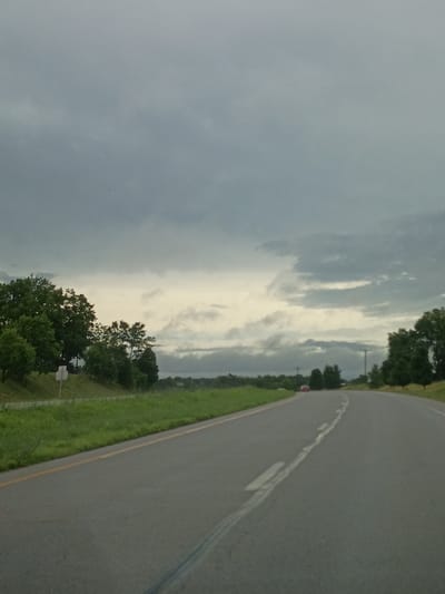 The image depicts a road in a rural or suburban area, likely in the city of Wilmore, United States of America. The road is paved and curves to the right, with a yellow line marking the edge of the lane. The sky is overcast and cloudy, suggesting a somber or potentially rainy day. Lush green grass and trees line the sides of the road, indicating a natural environment. In the distance, a red car is visible on the road, suggesting movement and travel. The scene evokes a sense of solitude and quietude, with the natural landscape dominating the view. There are no people clearly visible in the image, emphasizing the focus on the road and its surroundings.