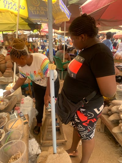 The scene depicts an outdoor market in Otuocha, Nigeria, under bright daylight, partially shaded by multiple large, colorful market umbrellas, including a yellow "GRAND MALL" branded umbrella. In the foreground, two women are present. The woman on the left, with blonde braided hair adorned with a pearl headband, wears a white t-shirt with a rainbow graphic and dark pants, and black sandals. She is leaning forward, actively examining or arranging various goods displayed on a wooden stall, which includes natural products like roots, powders in clear containers, and packaged items. To the right, another woman with dark braided hair (cornrows) wears a black t-shirt with a red, white, and blue "TOMMY S" logo, black, white, and red patterned shorts, and carries a black crossbody bag. She stands barefoot, facing generally towards the left woman and the stall. A light-colored pole, potentially supporting an umbrella or stall, stands centrally, anchored in a burlap sack. The stall to the right displays large, light-colored root vegetables. In the background, other market stalls, people, and a green plastic chair with a seated individual are visible, indicating a busy market environment with a dirt ground.