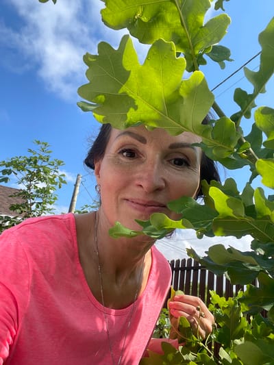 A woman with dark hair, wearing a pink t-shirt and a silver necklace, smiles as she peeks through green leaves. The background shows a blue sky with white clouds, a wooden fence, a tree, a wooden pole, and a portion of a house. The scene is outdoors in the city of Solov'evka, Russia.