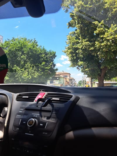 The image shows a view from inside a car, looking forward through the windshield. The dashboard of the car is visible in the foreground, with a car stereo and air vents. Outside the car, there is a street scene in the city of Popovo, Bulgaria. Trees line the street, and buildings are visible on either side. The sky is blue with some clouds. The overall impression is that of a sunny day in a small town.
